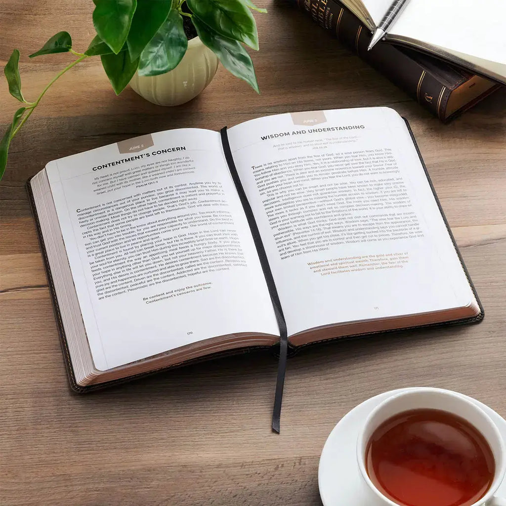 An open book with printed pages spread open on a wooden table, styled with a small plant, a cup of tea, and a dark object in the background.